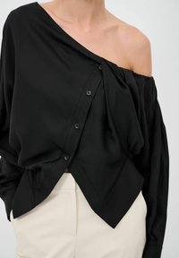 Camicia nera con bottoni a maniche lunghe e design off-shoulder, caratterizzata da una texture liscia e un orlo frontale piegato distintivo.