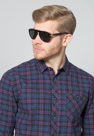 Occhiali da sole con lenti scure e montatura tartarugata. Indossati con una camicia a maniche lunghe a quadri nei colori viola e blu.
