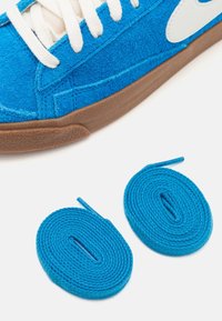 Baskets en daim bleu avec lacets blancs et semelle marron à côté de deux lacets bleus enroulés sur fond blanc.