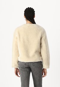 Giacca in finta pelliccia di colore beige chiaro, con una texture morbida, colletto rotondo e spalle scese. Abbinata a jeans grigi a vita alta e con texture.