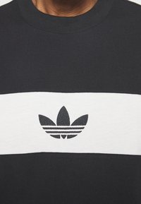 Černobílá mikina s kulatým výstřihem a horizontálním vzorem, s vyšitým logem Adidas trefoil uprostřed. Bavlněný materiál, regular fit.