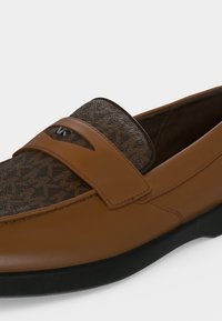 Καφέ loafer από δέρμα με επάνω μέρος από υφασμάτινο μοτίβο και μικρό ασημένιο λογότυπο "MK" στο λουράκι, με μαύρη σόλα.
