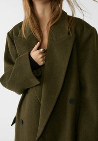 Dubbelzijdige olijfgroene jas van een gestructureerde wolmix, met brede revers en zwarte knopen. Gelaagd ontwerp onthult een zwarte top daaronder.