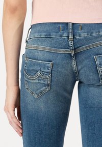 Blue denim jeans featuring a fitted design, back pockets with curved seams, and subtle fading for texture. Stitched accents in contrasting thread.