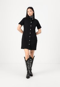 Vestido de ganga preto, com botões e mangas curtas, com um corte estruturado. Botões prateados em destaque e botas de cowboy em contraste com bordados brancos.