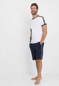 Weißes Baumwoll-T-Shirt mit marinefarbenen Akzenten und Schulterdetails, kombiniert mit marineblauen Shorts mit seitlichen Streifen. Model steht barfuß.