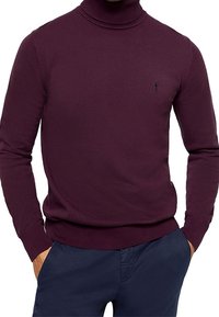 Homme portant un pull à col roulé ajusté bordeaux avec un petit logo noir brodé sur la poitrine, porté avec un pantalon bleu marine.