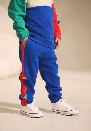 Kind trägt blaue Jogginghose mit bunten dreieckigen Spitzen entlang der Seitennähte und weißen Turnschuhen, Hände in den Taschen.