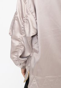 Primo piano della manica di un bomber in raso beige chiaro con dettaglio elastico arricciato e cerniera dorata sull'orlo laterale, indossato da una persona.