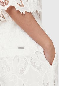 Valkoiset pitsishortsit, joissa on monimutkaisia kukkakuoseja, teksturoitu pinta ja hopeiset metalliosat takataskussa.