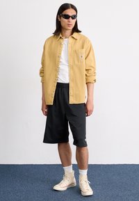 Gul denim skjorte, hvid t-shirt, sorte shorts og beige høj-top sneakers. Model iført sorte solbriller, stående på en blå tæppe baggrund.
