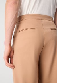 Los pantalones beige tienen una textura suave, una cintura de altura media y dos bolsillos traseros. La tela parece ligera y fluida.