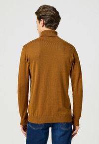 Brązowy sweter z wysokim kołnierzem, prążkowanymi mankietami i dołem. Gładka faktura i dopasowany krój, noszony na niebieskich dżinsach.