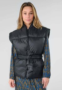 Veste noire matelassée avec un col haut, une taille cintrée par un lien, et des épaules larges. Portée sur une robe à motifs présentant des tons bleus et bruns.