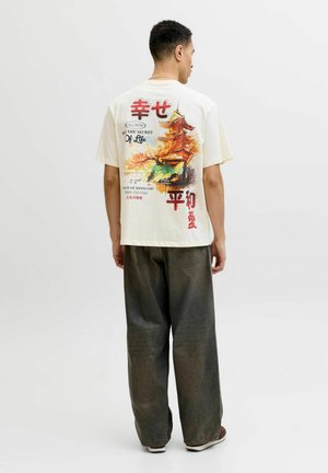 Mężczyzna odwrócony tyłem, ubrany w beżową koszulkę z pomarańczową pagodą i japońskim napisem, w połączeniu z luźnymi ciemnymi spodniami i białymi trampkami.
