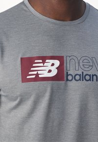Graues Baumwoll-T-Shirt mit einem burgunderroten rechteckigen Logo, darauf weißer "nb" Text und "new balance" in Marineblau, mit rundem Ausschnitt und weicher Textur.