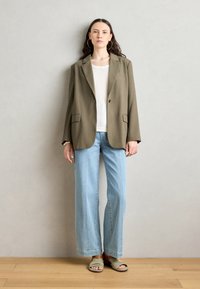 Blazer a medida verde oliva con solapas anchas, dos bolsillos frontales; combinado con una blusa blanca, vaqueros de pierna ancha azul claro y sandalias verdes.