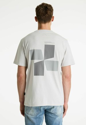 Férfi áll háttal, világosszürke pólót visel, amelyen absztrakt négyzet alakú szürke geometrikus minta és világoskék farmernadrág van.