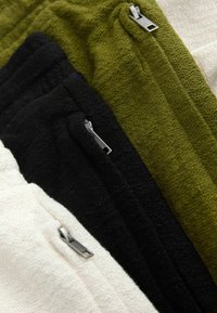 Gros plan sur trois pantalons en tissu texturé de couleur crème, noir et vert olive, chacun avec une fermeture éclair métallique visible sur la poche.