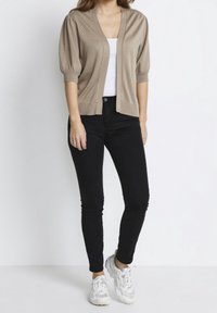 Beige stickad cardigan med korta puffärmar, öppen framsida, bärs över en vit topp och matchas med svarta jeans och vita sneakers.