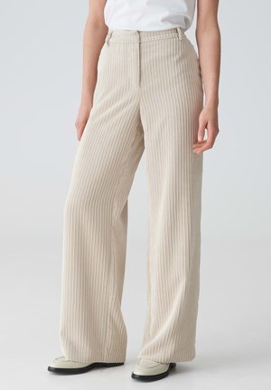 Beige wide-leg trousers with vertical ribbed texture, high waist, and side pockets, paired with light-colored shoes.