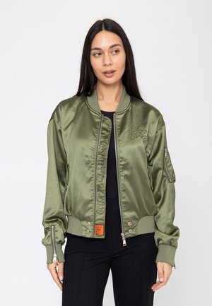 Vrouw met lang donker haar die een olijfgroene satijnen bomberjas draagt over zwarte kleding, staand tegen een effen witte achtergrond.