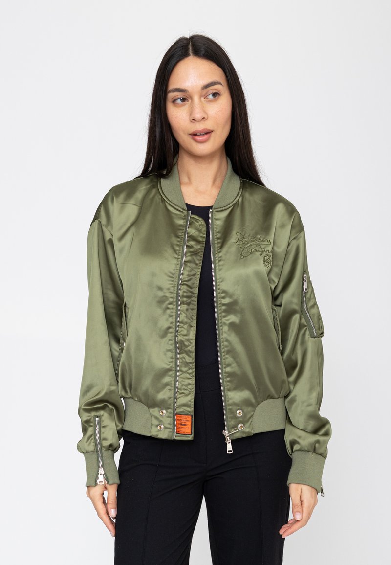 Femme aux longs cheveux foncés portant une veste bomber en satin vert olive sur des vêtements noirs, debout devant un fond blanc uni.