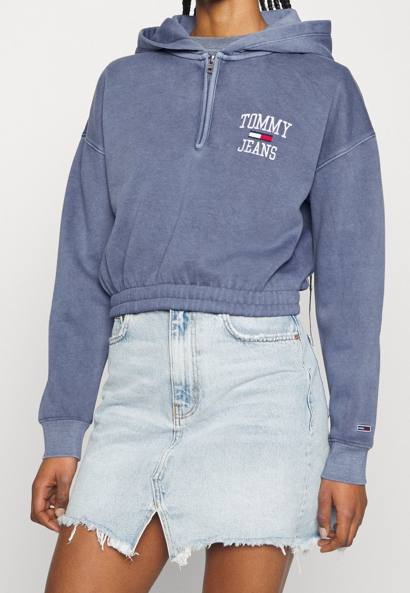 Personne portant un hoodie court bleu Tommy Jeans avec une fermeture éclair au quart et une jupe en denim bleu clair effilochée avec une fente à l'avant.