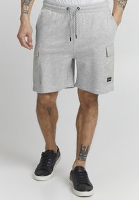 Shorts en coton gris avec taille élastique, cordon de serrage et deux poches cargo sur les côtés. Associé à des baskets noires. Présence de tatouages sur les jambes.