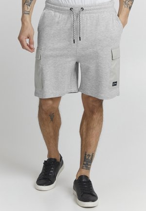 Graue Baumwollshorts mit elastischem Bund, Kordelzug und zwei seitlichen Cargo-Taschen. Getragen mit schwarzen Turnschuhen. Auf den Beinen sind Tattoos zu sehen.