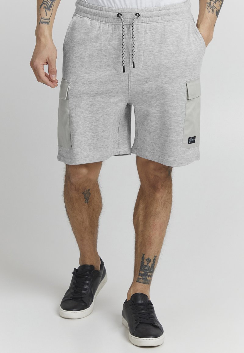 Pantaloni corti in cotone grigi con elastico in vita, cordoncino e due tasche laterali a soffietto. Abbinate a scarpe da ginnastica nere. Presentano tatuaggi sulle gambe.