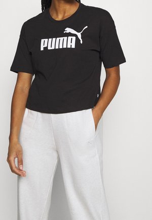 Persoon die een zwart Puma-t-shirt en lichtgrijze joggingbroek draagt, met één hand in de zak en de andere ontspannen langs het lichaam.