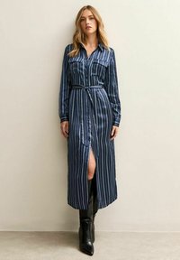 Kobieta w granatowej, pasiasto wzorzystej sukience midi z długim rękawem, z paskiem w talii i rozcięciem z przodu, w połączeniu z czarnymi kozakami za kolano.