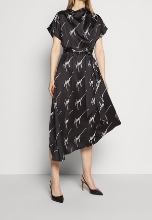 Žena v černých midi šatech s bílým nápisem "Hugo", krátké rukávy, pásek v pase a černé lodičky s špičatou špičkou.