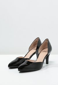 Escarpins en cuir verni noir avec un bout pointu et un talon aiguille de 7,5 cm. Les côtés ouverts révèlent une semelle intérieure nude avec une marque.