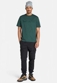 Vihreä puuvillainen T-paita, jossa kohokuvioitu "Timberland"-logo, yhdistettynä mustiin cargo-housuihin ja ruskeisiin vaelluskengiin. Harmaa pipo viimeistelee lookin.