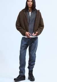 Veste zippée brune en peluche, chemise à rayures avec boutons, t-shirt blanc en dessous, pantalon gris ample, bottes à lacets noires, tenue décontractée.