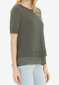 Olivengrøn kortærmet top med lagdelt design, featuring ribbet kant og struktureret nederste sektion, parret med lyseblå slidte jeans.
