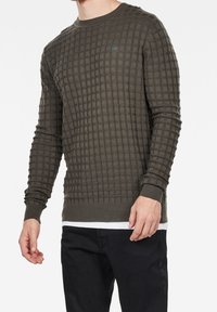 Sweter w oliwkowym zielonym kolorze z teksturą, z wzorem w kratkę, okrągłym dekoltem, długimi rękawami oraz ściągaczami na rękawach i u dołu. Nosi się go na białej koszuli.