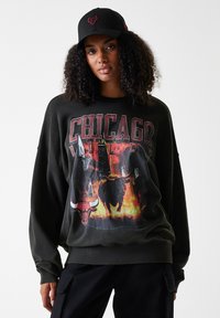 Sweatshirt noir surdimensionné présentant un graphisme des Chicago Bulls avec des accents rouges et jaunes, sur fond de ville enflammée.