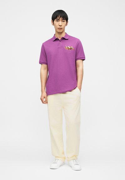 Άνδρας με μωβ πόλο μπλούζα με κεντημένους παίκτες πόλο, κρεμ παντελόνι και λευκά αθλητικά παπούτσια, στέκεται μπροστά σε απλό λευκό φόντο.