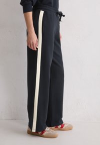 Person trägt schwarze weite Hosen mit einem weißen Seitestreifen und beigen Sneakern mit roten Akzenten und steht vor einer hellen Wand.