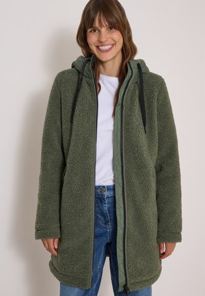 Femme souriante portant une veste à capuche en polaire vert olive par-dessus une chemise blanche et un jean bleu sur un fond clair uni.