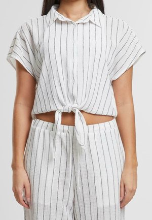 Femme portant une chemise blanche à manches courtes boutonnée et nouée à la taille, ainsi qu'un pantalon taille haute assorti avec des rayures verticales noires.