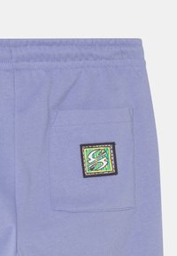 Calças de treino lilás claras com cintura elástica, apresentando um bolso quadrado decorado com um patch colorido que exibe um "S" estilizado.