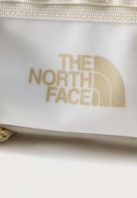 Το λογότυπο της The North Face σε μπεζ χρώμα πάνω σε μια υφασμάτινη επιφάνεια με υφή και ανοιχτόχρωμο φόντο, με μια μπεζ λουρίδα από πάνω.
