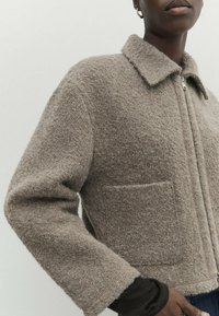 Mörkhyad person som bär en strukturerad beige zip-up jacka med krage och bröstficka, kombinerad med mörka innersleeves och blå jeans.