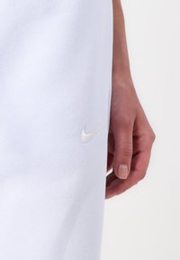 White Nike sweatpants with a smooth texture, featuring a small embroidered silver logo on the left leg, hand partially visible.