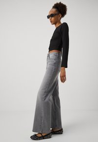 Donna con i capelli ricci raccolti in una coda di cavallo, che indossa occhiali da sole neri, un top lungo a maniche nere, jeans grigi a gamba larga e ballerine nere con borchie, in piedi di profilo.