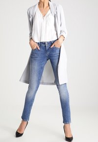Jasnoszary, długi żakiet na białej bluzce, w zestawie z niebieskimi dopasowanymi dżinsami i czarnymi szpilkami. Gładki materiał i dopasowany krój.
